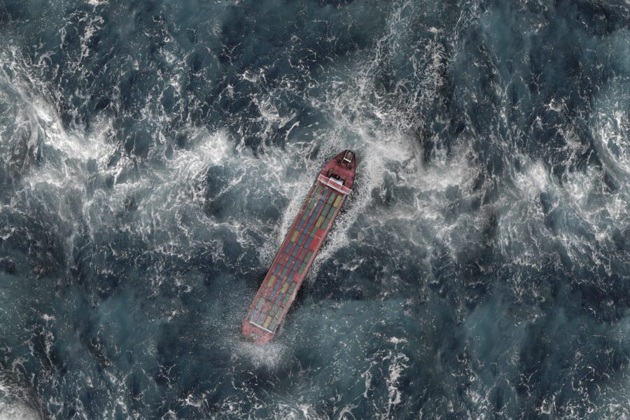 Supply chain resilience: aerial view of a container ship navigating rough, foamy seas during a global disruption.